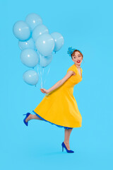 Brightly dressed woman in yellow dress joyfully holding blue balloons against a solid blue background