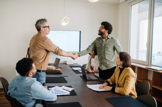 Two businessmen shake hands at the end of a successful business meeting. A diverse group of colleagues watches the interaction in a modern conference room.