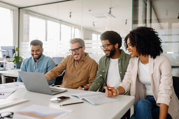 A diverse group of professionals laughs together while working on a laptop in a modern office. The team is engaged in a business discussion, showcasing positive teamwork and collaboration.