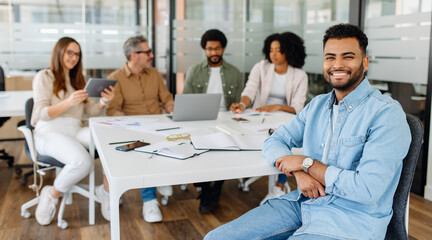 Fototapeta premium Young professional in denim shirt smiling confidently while sitting at a business meeting with a diverse team in a modern office space.