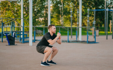 A man exhibits focus and strength as he performs squats in a sunny outdoor gym, dressed in sporty...