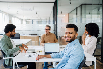 A diverse group of professionals engaged in a team meeting in a modern office. A senior executive leads the discussion while colleagues actively participate, taking notes and working on laptops