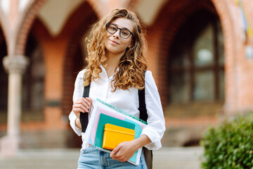 Beautiful student with curly hair stands with a stack of books on the background of a college...