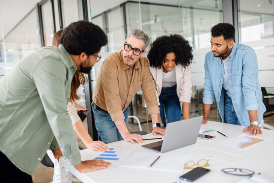 Team of professionals gathered around a laptop, analyzing data and discussing strategies in a modern office. A diverse group collaborating on a business project with documents, graphs, and technology