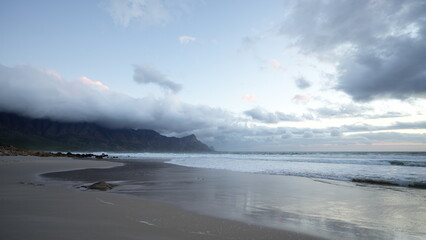 Waves gently lap against the sandy shore as dusk settles over the beach, casting a soft light on the distant mountains shrouded in clouds. It's a peaceful moment by the sea.