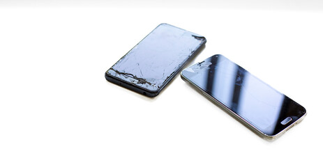 Two damaged smartphones with cracked screens stacked on a white surface, reflecting light. Concept of device repair, damage, and technology issues