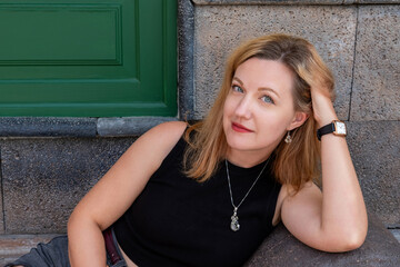 Confident blonde woman with deep blue eyes wearing a black sleeveless top, a sliver pendant necklace, elegant drop earrings posing casually against a stone bench with one arm resting behind her head