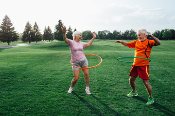 Two older individuals are joyfully exercising outdoors with hula hoops, showcasing energy and fun in a green park setting during daylight © Peakstock