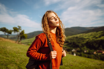 A young woman with a backpack is walking on a green hill. A female traveler in a hat is enjoying...