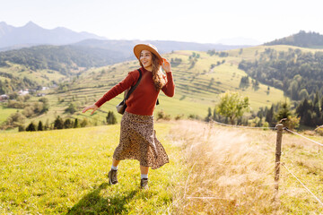 A young woman with a backpack is walking on a green hill. A female traveler in a hat is enjoying...