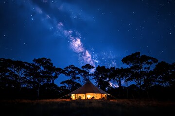 a glowing tent under a breathtaking starry sky. The Milky Way is visible above, with silhouettes of trees 
