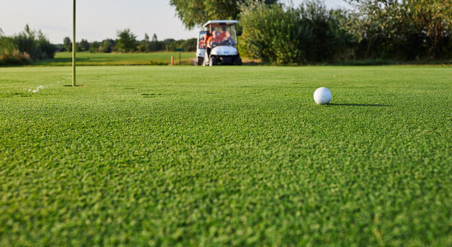 A golf ball rolls towards the hole on a vibrant green course while a golf cart waits nearby in the warm afternoon sunlight