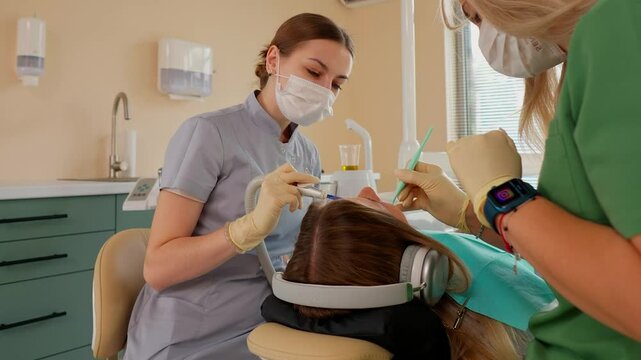 Blonde Woman Dentist with Assistant Treat Teeth of Female Patient with Special Instrument. Treatment of Caries and Plaque in Dental Clinic. Concept of Medicine and Healthcare. Slow Motion.