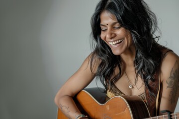 Portrait of a grinning indian woman in her 30s playing the guitar in soft gray background