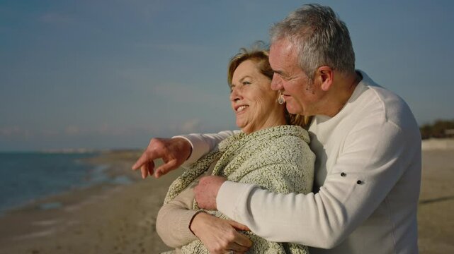 Happy  family elderly husband cover his wife with a blanket and hugging each other on tropical beach at summer sunset. Retirement senior couple enjoy romantic outdoor lifestyle vacation together.