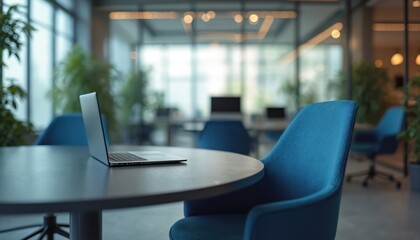 Laptop on table in blurred modern open office room with plants, soft lighting. Sleek design business workspace with blue armchairs. Contemporary professional corporate workplace ready for use.