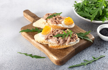 Open toast with fresh bruschetta, canned tuna, boiled egg and arugula on a wooden board. Top view.