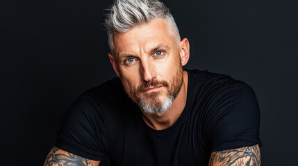 Fototapeta premium A man with light gray hair and a well-groomed beard showcases his vibrant blue eyes and extensive tattoos while posing confidently against a dark backdrop in a studio