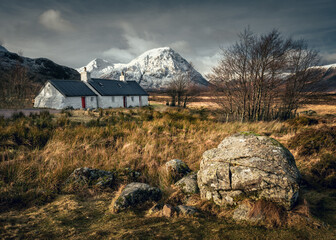 View of typical Scottish stone house with moor in the foreground and the snow capped mountain in the background with dramatic atmosphere during winter day, Glencoe valley, Scottish Highlands, Scotland © Ji