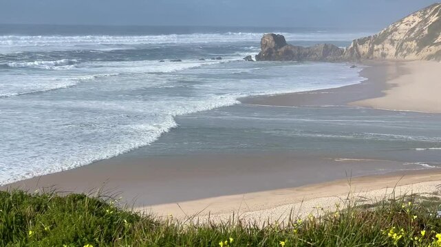 Ocean waves crash onto the sandy shore of Praia de Paredes da Vit&oacute;ria, with rocky cliffs in the background. The shimmering water and coastal breeze. Green grass and yellow flowers frame the view.