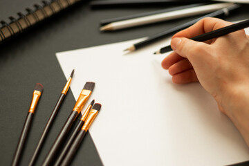 A monochrome art-themed photo featuring graphite pencils and metallic paints on a black background,...
