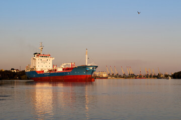 A large cargo ship floats on calm waters near a busy port with towering cranes in Ukraine