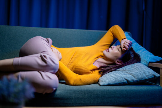 Young woman lying on the sofa at night, holding her belly and head, suffering from abdominal pain and headache