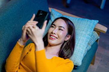 Young woman relaxing on cozy sofa, scrolling smartphone with joyful smile during late evening...