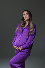 Portrait of attractive pregnant woman in violet costume, trousers, studio photoshoot. Fashion photo of pregnancy. Young pretty girl with long hair touching her belly. Isolated, grey background