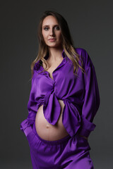 Portrait of attractive pregnant woman in violet costume, trousers, studio photoshoot. Fashion photo of pregnancy. Young pretty girl with long hair touching her belly. Isolated, grey background