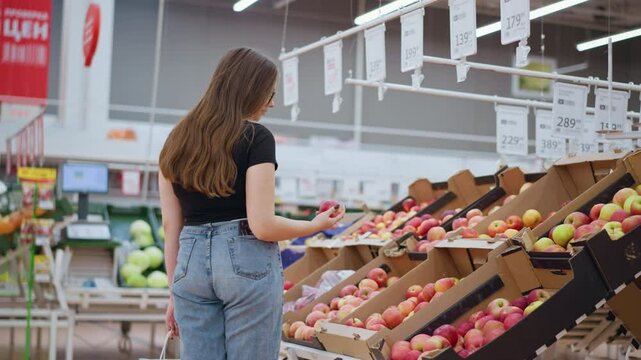 Rear view of lady with phone in back pocket picking red apple from display, observing it, and dropping it back, price tags and signs visible in the background of bright store environment