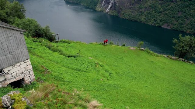 Aerial Drone Over Turf Houses Towards Wide Fjord Geiranger Norway Seven Sisters Waterfall. Photographer Couple in Orange Jackets on Edge