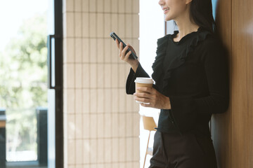 Relaxing Moment: A woman stands casually, engrossed in her phone while holding a cup of coffee by a...