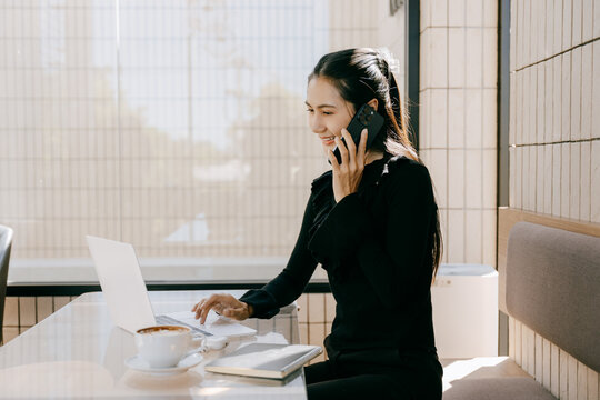 Busy businesswoman working on laptop: A modern businesswoman, engrossed in her work, engages in a phone call while typing on her laptop at a bright, contemporary cafe setting.
