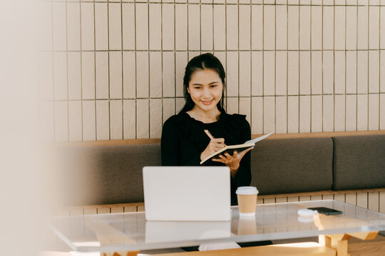 Focused Productivity: A young professional, immersed in her work, smiles brightly while making notes in her journal and working on a laptop in a cozy and inspiring environment. - Powered by Adobe