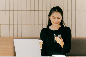 Digital Engagement: An Asian woman, engrossed in her phone, balances a coffee cup with a laptop...