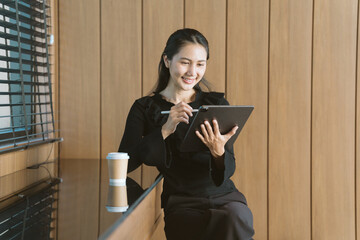 Digital Age Efficiency: A smiling Asian businesswoman leans on a sleek wooden counter, engrossed in...