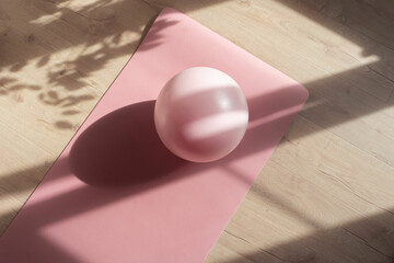 Flat lay of fitness ball for pilates workout on pink yoga mat with shadows and sunlight which convey the atmosphere of early morning