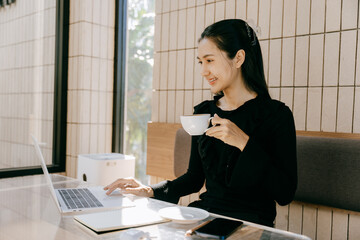 Digital Nomad with Morning Coffee: A smiling woman engrossed in her laptop while sipping from a...