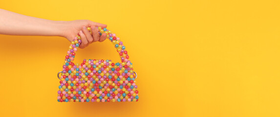 Banner with cute multicolored handbag made from acrylic beads in a female hand in front of yellow background.