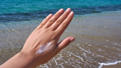 Woman’s hand with sunscreen cream against a turquoise sea backdrop, emphasizing summer skincare...