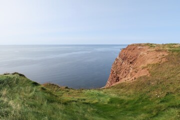 Vom Klippenrandweg auf der deutschen Hochseeinsel Helgoland sind die Felsen aus Buntsandstein, Gras darauf und die Nordsee unten zu sehen. Die See ist sehr ruhig an diesem Sommertag.