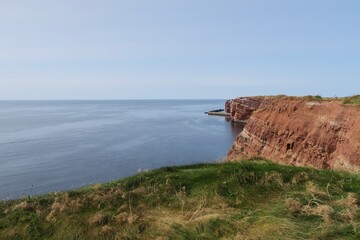 Vom Klippenrandweg auf dem Oberland der deutschen Hochseeinsel Helgoland sind die Felsen aus Buntsandstein, Gras darauf und die Nordsee unten zu sehen. Die See ist sehr ruhig an diesem Sommertag.