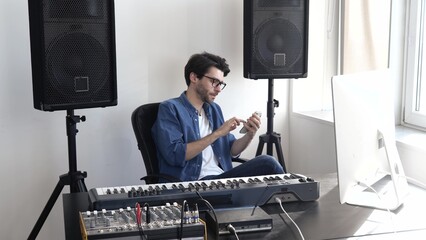Young man in sound recording studio. Sitting at keyboard and typing on smartphone. Texting or...
