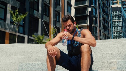 Young man exercising outside. Tired exhausted guy sitting on steps on street and drink water....