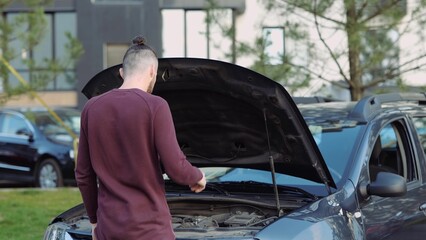 Young man outside car. Open its hood and helpless with broken engine. Calling on phone to get help...