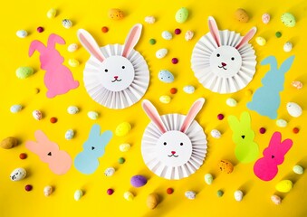 Spring, Easter holiday, on a yellow background are cute, funny Easter paper bunnies, colored candies and bright Easter eggs. Flat lay, top view. The concept of children's creativity, handmade
