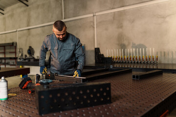 Metalworker wearing gloves and using a ruler to measure a metal frame on a welding table in a...