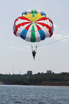 A person parasailing high above the water with a colorful parachute featuring red, blue, green, yellow, and white panels. The silhouette of the parasailer contrasts against the bright sky
