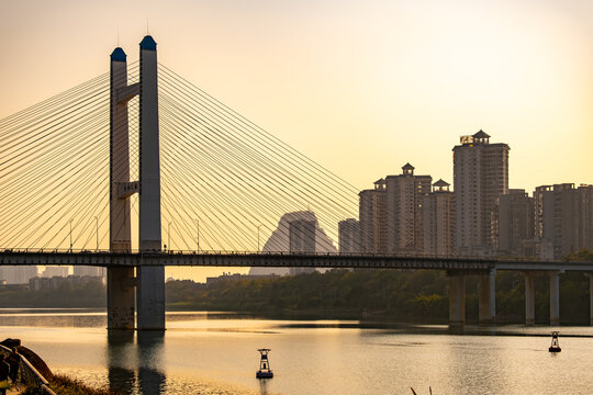 Huxi Bridge in Liuzhou, China, the cable-stayed bridge's twin towers and elegant suspension cables silhouetted against a golden sunset sky, with the city's modern skyline visible in the background. 